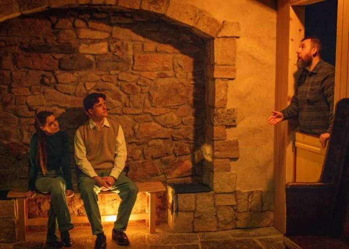 Visitors seated by a glowing stone hearth as a storyteller speaks from the doorway in the National Leprechaun Museum’s cottage scene.