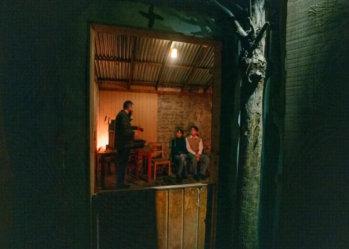 Storyteller speaking to visitors inside a softly lit cottage scene at the National Leprechaun Museum, viewed through a rustic doorway.