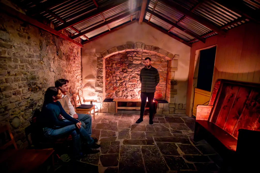 Guide sharing Irish folklore with two visitors inside a softly lit stone cottage room at the National Leprechaun Museum.