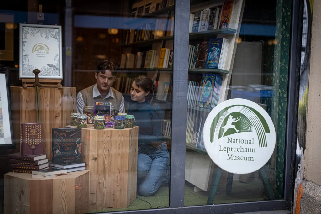 Two visitors browsing folklore books and gifts inside the National Leprechaun Museum shop, viewed through the window with the museum logo displayed.