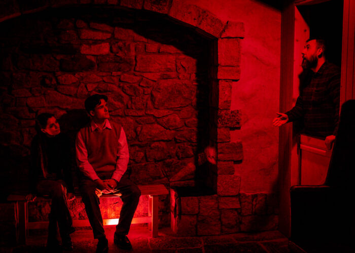 A storyteller stands in the doorway of a rustic cottage, speaking to two museum guests sitting down nearby. The scene is illuminated with a stark red light.