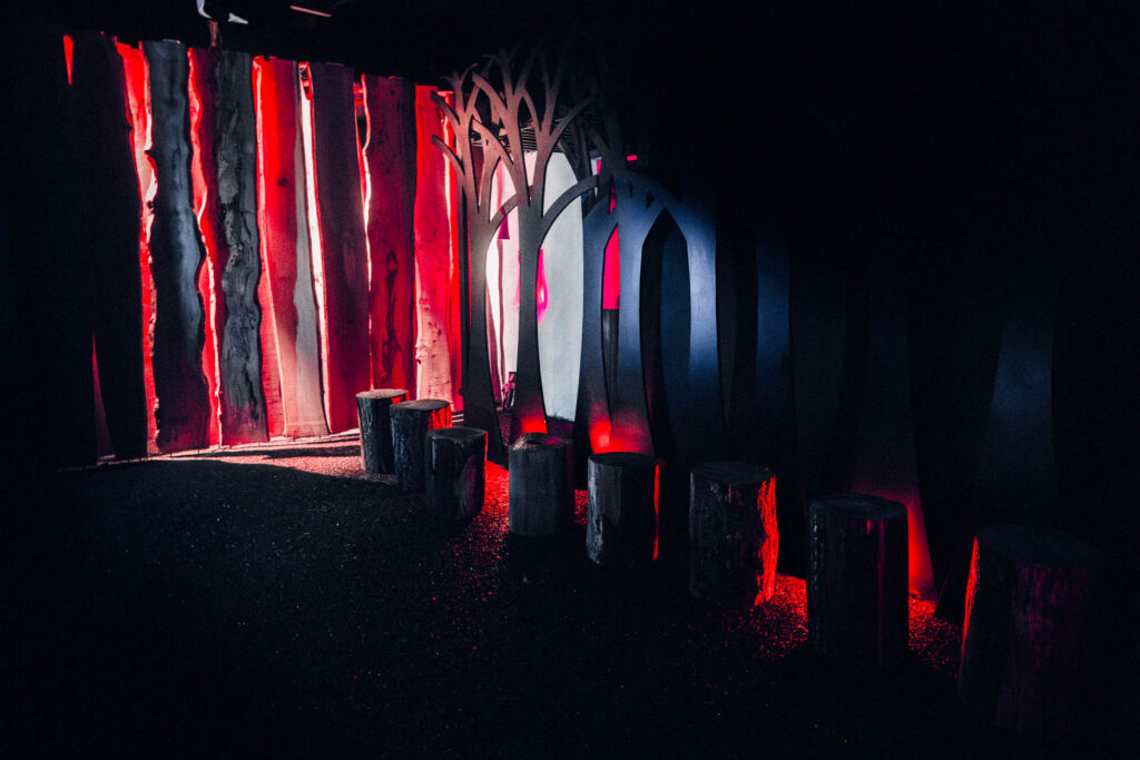 A forest-like scene in the museum interior, with logs for seating and flat, tree-shaped decorations behind them. The image is dark, illuminated with a narrow band of red light.