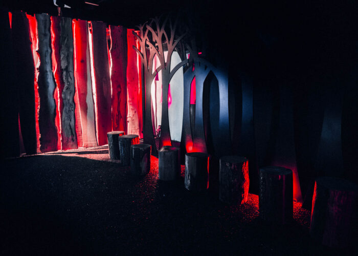 A forest-like scene in the museum interior, with logs for seating and flat, tree-shaped decorations behind them. The image is dark, illuminated with a narrow band of red light.