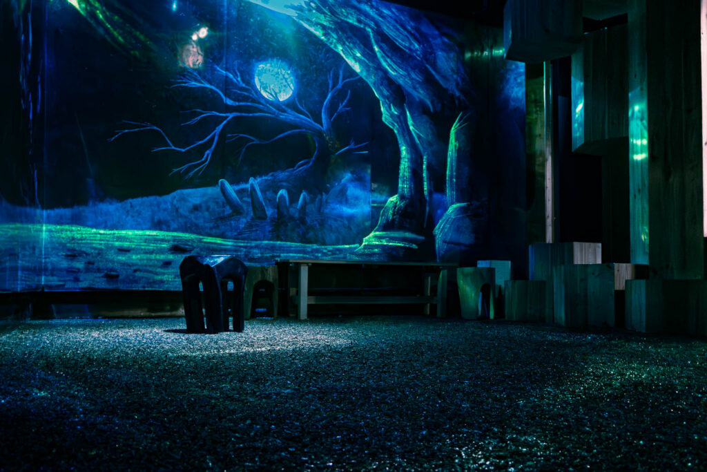 Seating in a room in the museum, tinted blue. In the background, a projection of a cave entrance can be seen, with a gnarled, leafless tree illuminated by a full moon.
