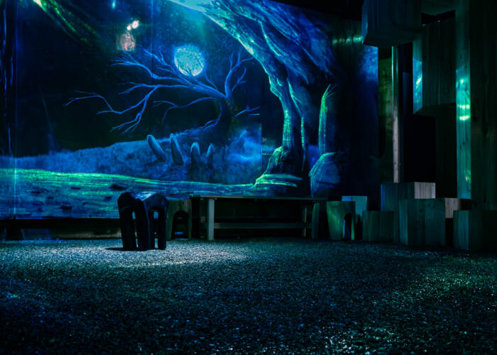 Seating in a room in the museum, tinted blue. In the background, a projection of a cave entrance can be seen, with a gnarled, leafless tree illuminated by a full moon.