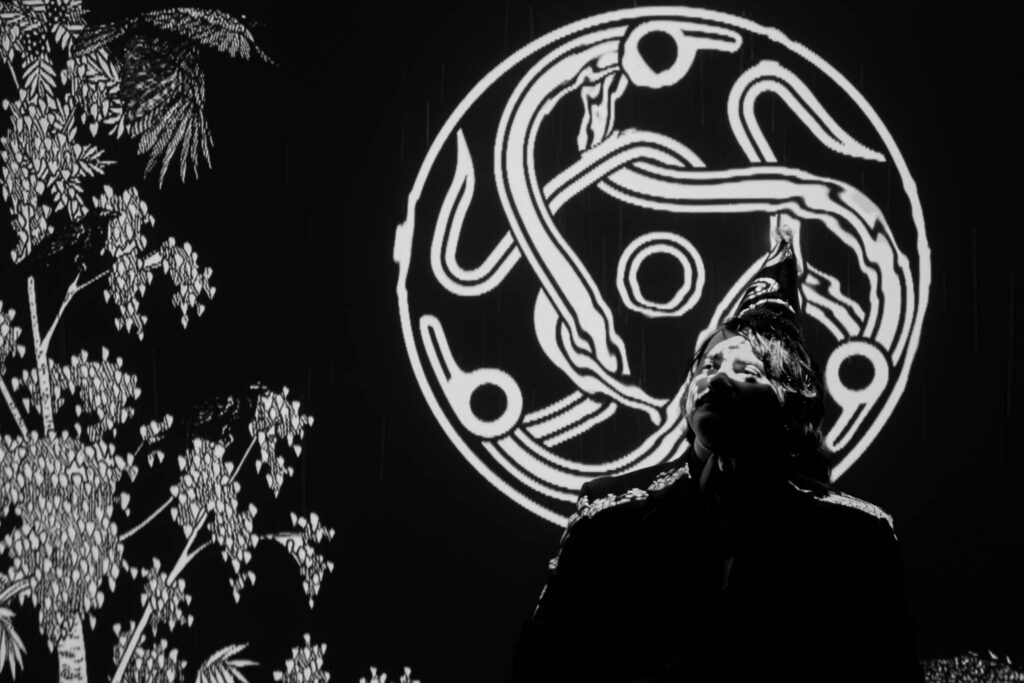 A black and white image of a storyteller, illuminated by the light of the Darkland logo, an intertwining celtic knot. To their left, illustrations of crows in branches can be seen.