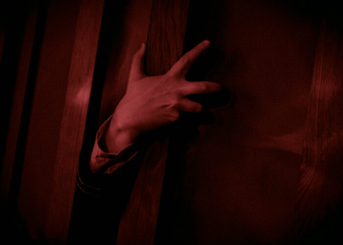 A hand reaching around a doorway. The entire image is tinted red.