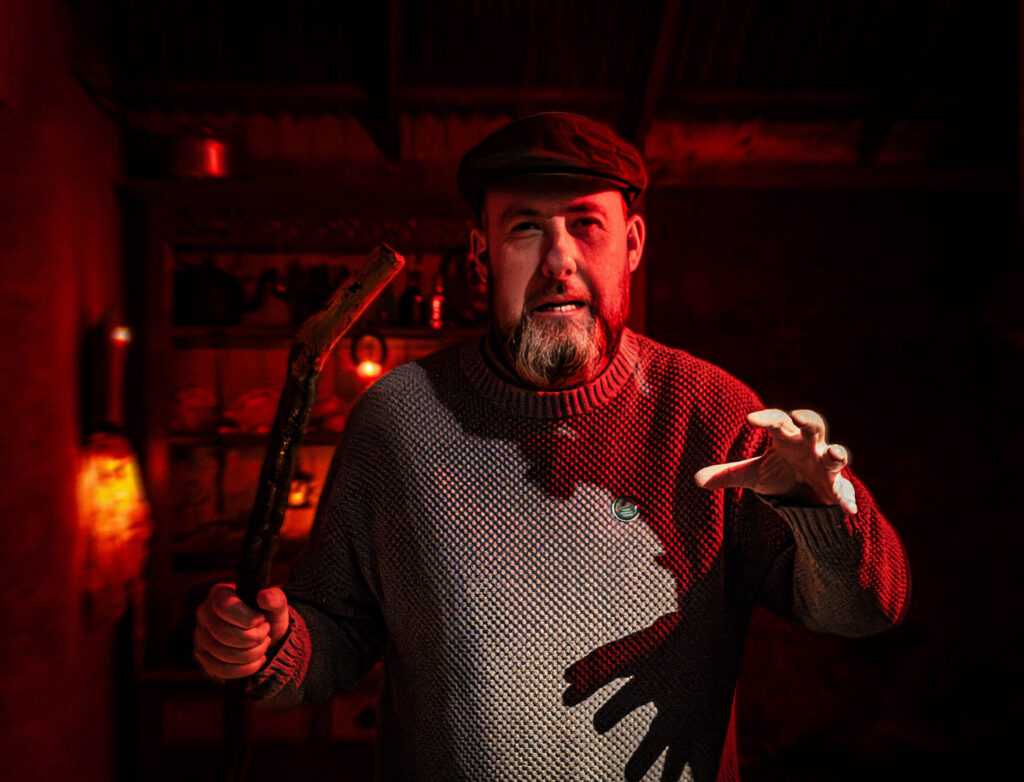 A Storyteller with a stick stands in the doorway to a dimly lit cottage, with one hand outstretched to the viewer. His face is illuminated by red light.