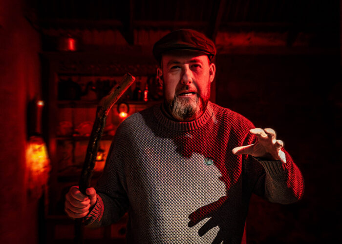 A Storyteller with a stick stands in the doorway to a dimly lit cottage, with one hand outstretched to the viewer. His face is illuminated by red light.