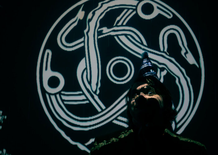 A storyteller staring into the camera, illuminated by the Darkland logo, a celtic knot of three serpentine figures intertwining.