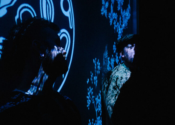 Two storytellers looking at eachother, one facing the camera, one facing away. One is illuminated with the Darkland Logo, the other by illustrations of bushes and crows.