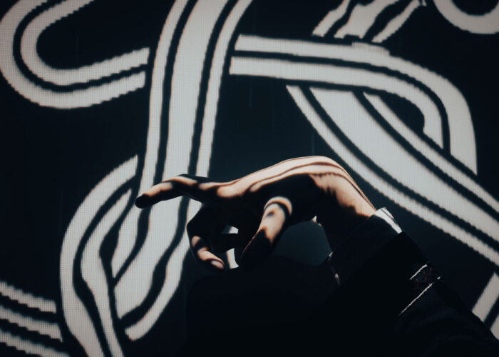 An outstretched hand in a clawed gesture is illuminated by a celtic spiral projected onto it and the background.