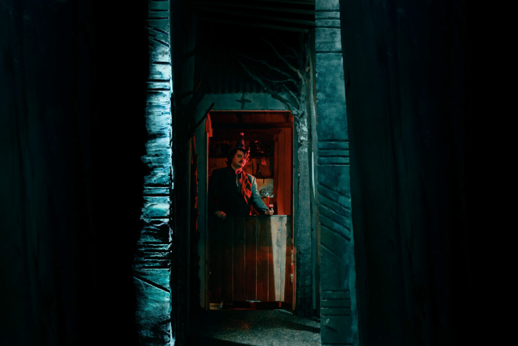 A storyteller, illuminated in red, stands in the doorway of a rustic cottage, flanked by two large pillars marked with Ogham writing.