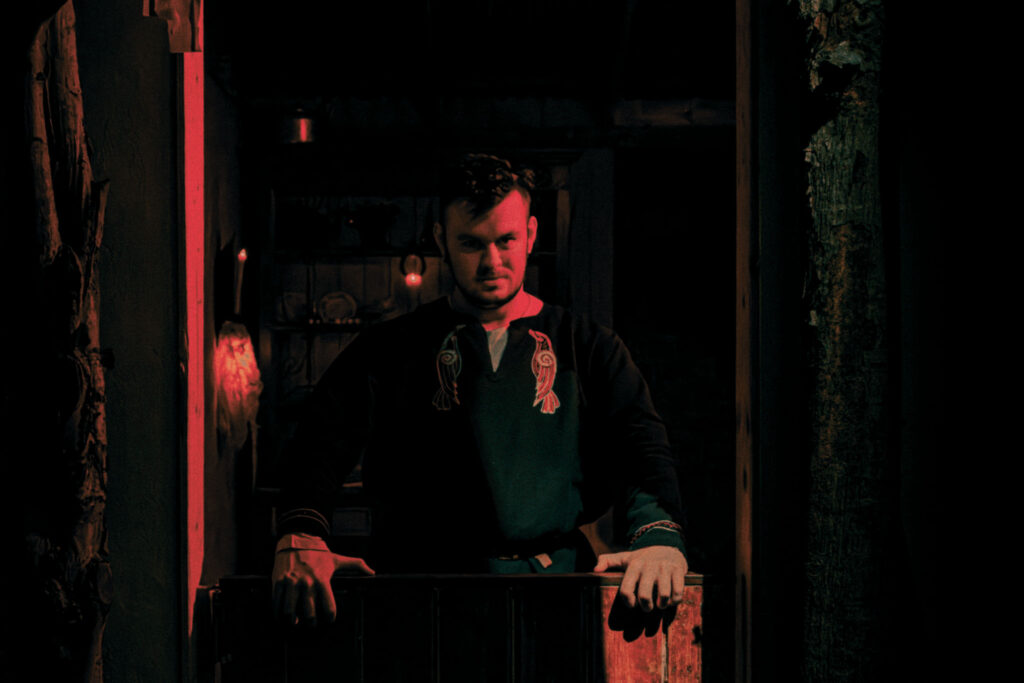 A storyteller, dressed in a viking-style tunic, stands in the doorway to a rustic cottage, illuminated by red light.