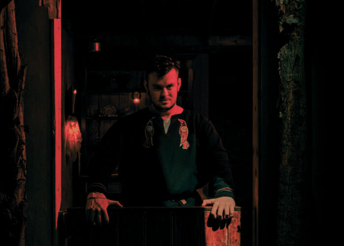 A storyteller, dressed in a viking-style tunic, stands in the doorway to a rustic cottage, illuminated by red light.