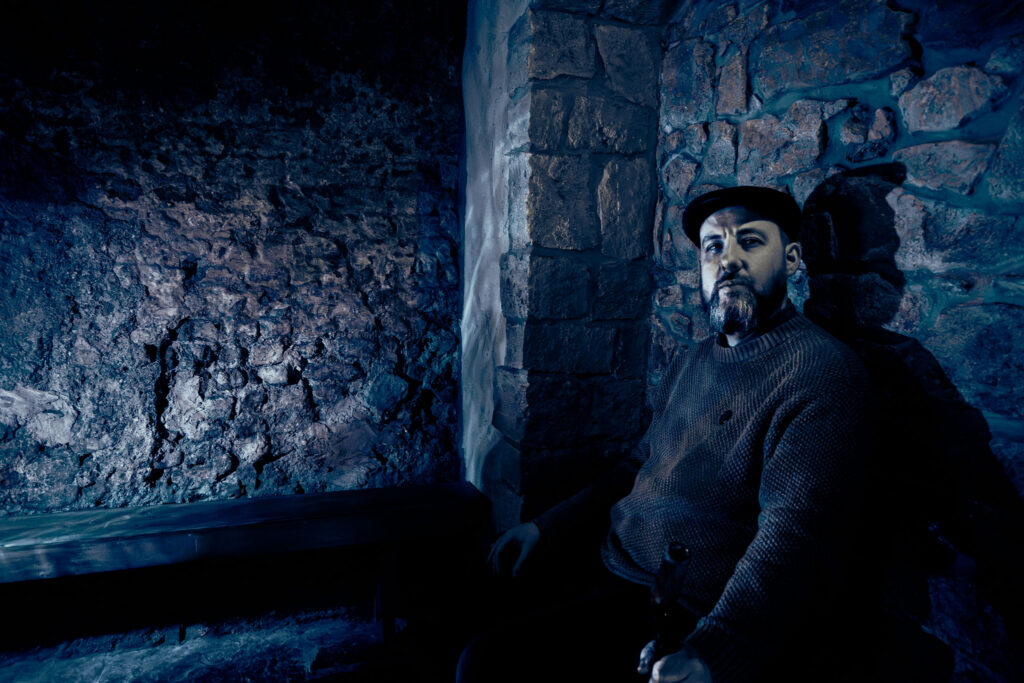 A storyteller sits in a rustic stone cottage, illuminated in blue light.