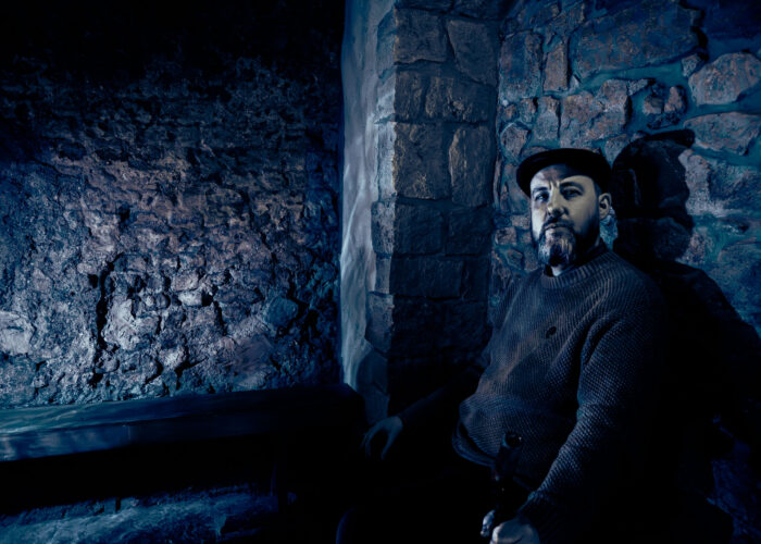 A storyteller sits in a rustic stone cottage, illuminated in blue light.