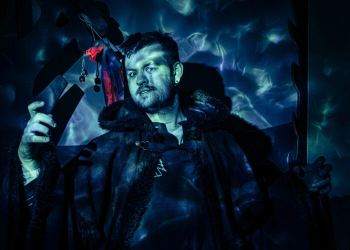 A storyteller wearing a dark cloak looks into the camera, illuminated by a blue and green water-caustics projection.