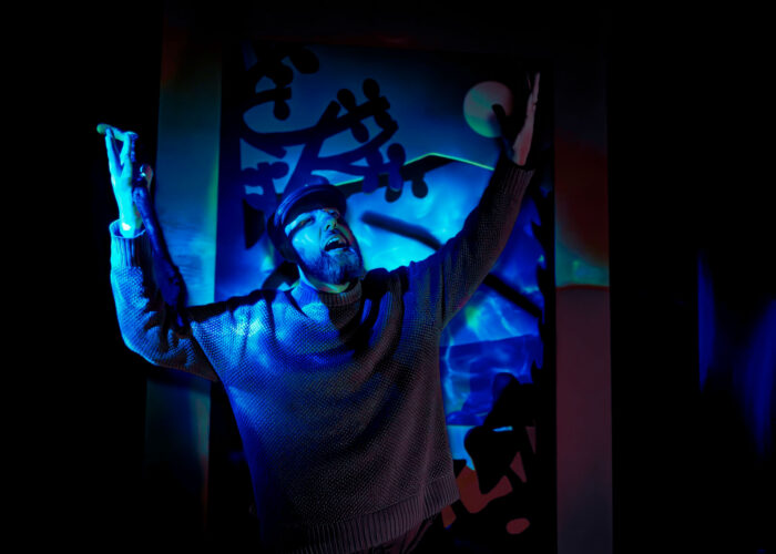 A storyteller standing in front of a forest diorama. He's illuminated by blue light, his arms raised up in the air as he yells.