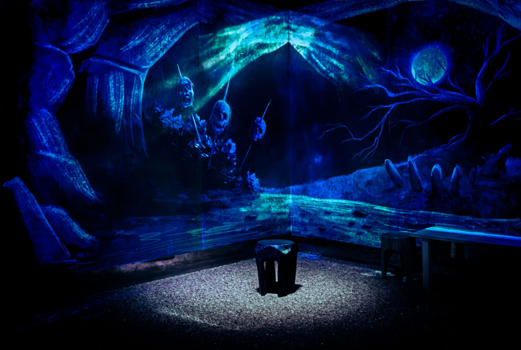 A single black stool in the middle of a blue room. Behind it, a cave-like background is projected. The projected scene is of three severed heads on spikes, with a large, leafless tree in the background surrounded by standing stones.