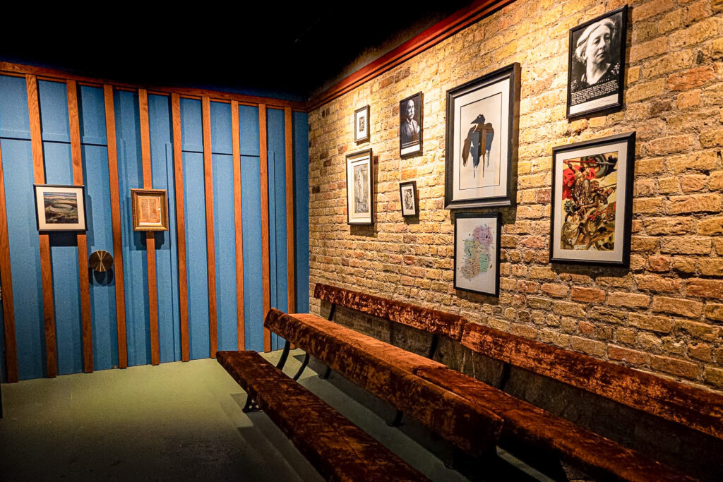 Two rows of benches sit in front of a brick wall. Mounted on the wall are eight photos and paintings of figures from Irish folklore. An adjoining blue wall can be seen, decorated with a series of thin wooden planks and two more pictures.