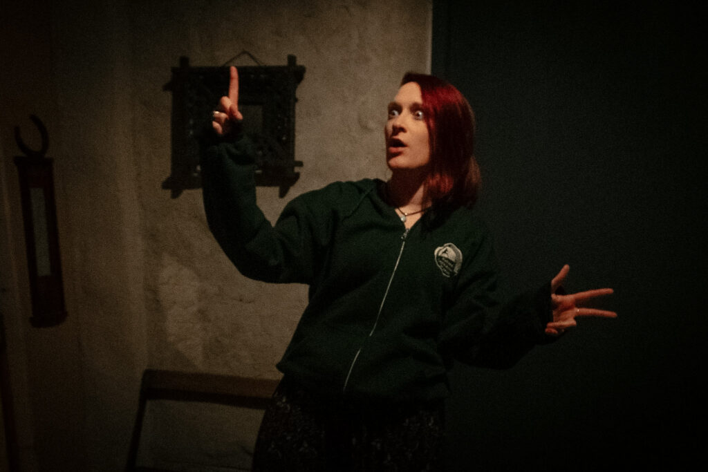A storyteller stands in a rustic cottage interior, talking with an animated expression. They wear a hoodie displaying the logo of the National Leprechaun Museum.