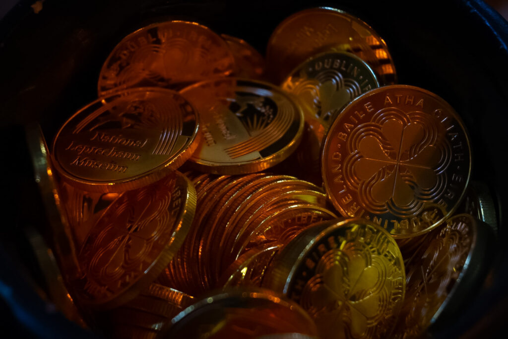 A pot of gold coins, each emblazoned with the National Leprechaun Museum logo or a four leafed clover.