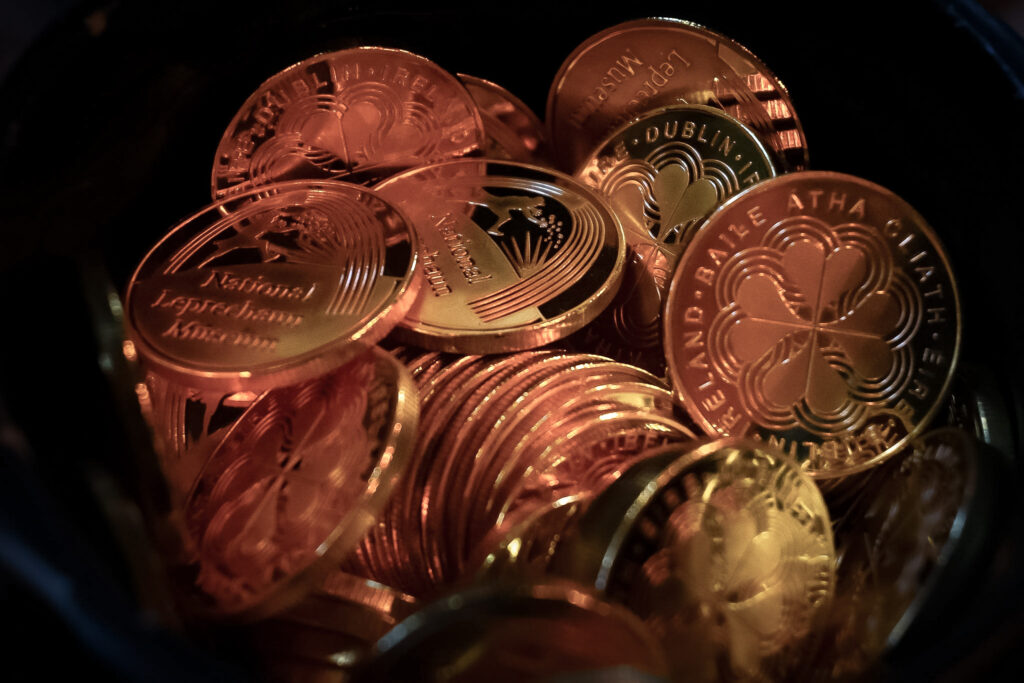 A pot of gold coins, each emblazoned with the National Leprechaun Museum logo or a four leafed clover.