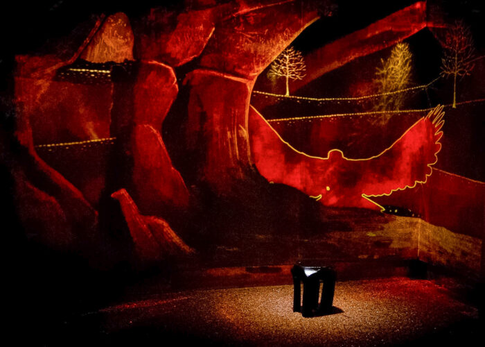 The Fairy Hill of the museum. A reddish background is projected onto the walls, showing a cave and an illustrated landscape scene. A spotlight illuminates a black stool in the foreground.