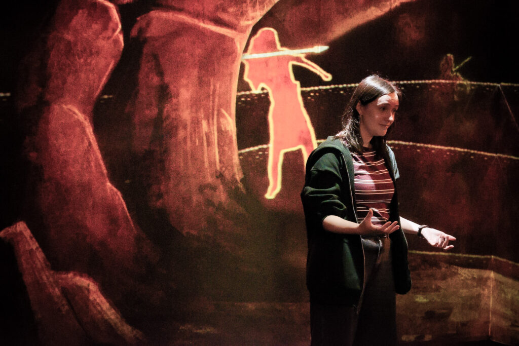 A storyteller stands in the Fairy Hill of the museum. She is illuminated by a spotlight. Behind her, an illustrated background is projected onto the walls.