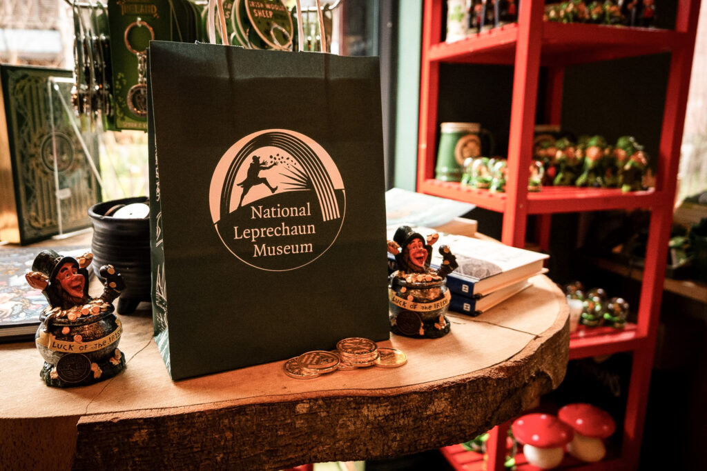 The shop of the museum. A green bag bearing the museum's logo sits on a table made from a tree trunk. Around it are books, coins, small leprechaun figurines and other souvenirs.
