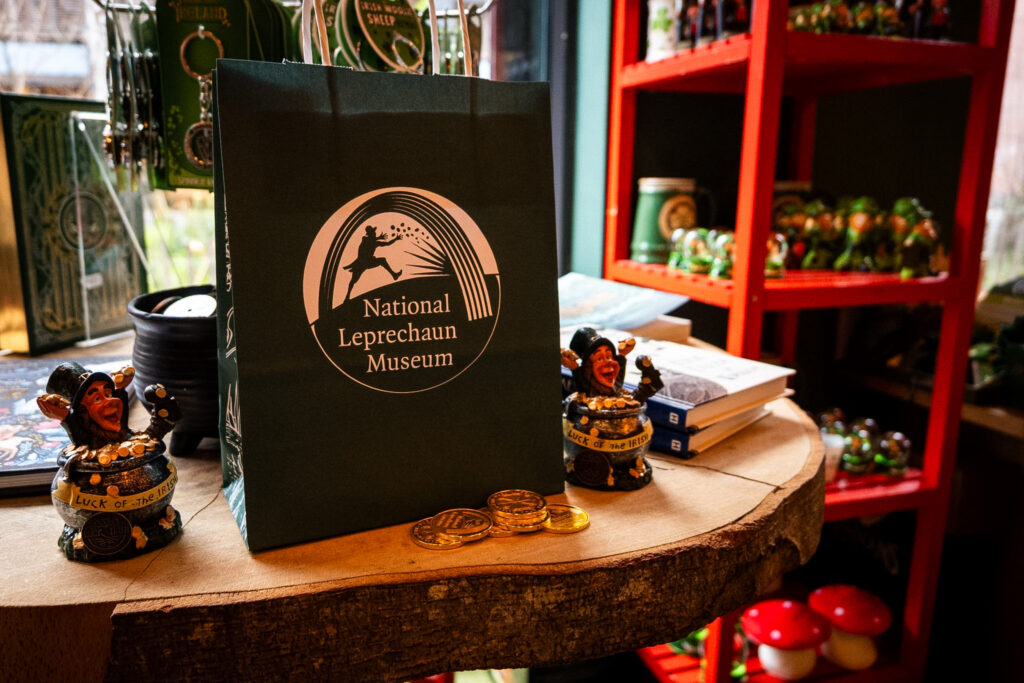 The gift shop of the museum. A green bag with the museum's logo is shown on a wooden table, surrounded by leprechaun figurines, books and other gifts.
