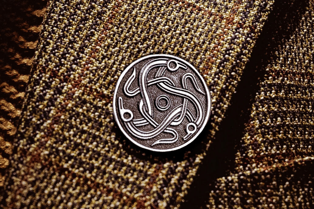 A metal pin badge of the darkland logo, three celtic figures intertwined together in a circle. The badge is mounted on tweed fabric.