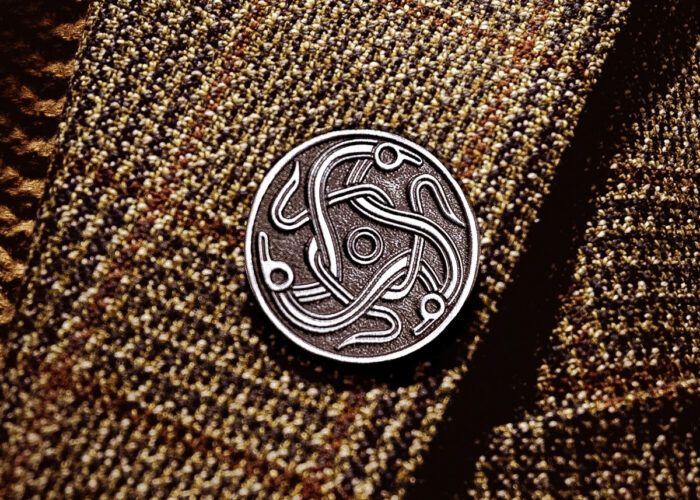 A metal pin badge of the darkland logo, three celtic figures intertwined together in a circle. The badge is mounted on tweed fabric.