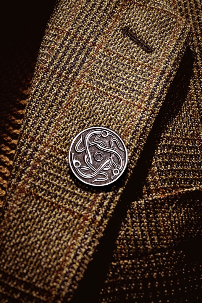 A silver pin badge of the darkland logo on the lapel of a tweed jacket.