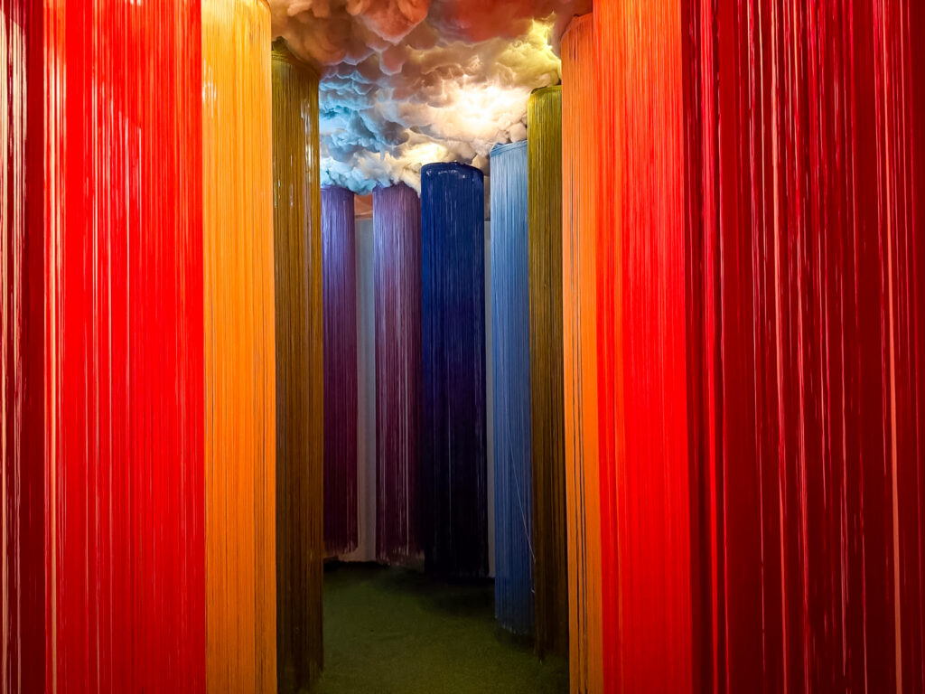 A rainbow-themed corridor, with long strings in the colours of the rainbow hanging from a cloud like ceiling.
