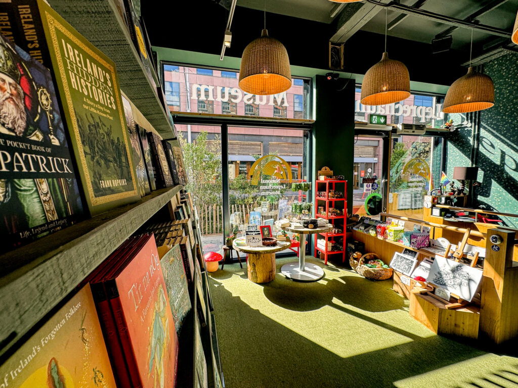 A view of the museum's gift shop, with sunlight shining in through large windows. A bookshelf containing books on Irish folklore is on the right, and to the left are tables and shelves displaying souvenirs and prints.