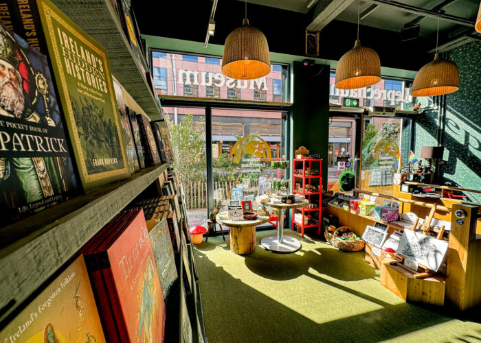 A view of the museum's gift shop, with sunlight shining in through large windows. A bookshelf containing books on Irish folklore is on the right, and to the left are tables and shelves displaying souvenirs and prints.