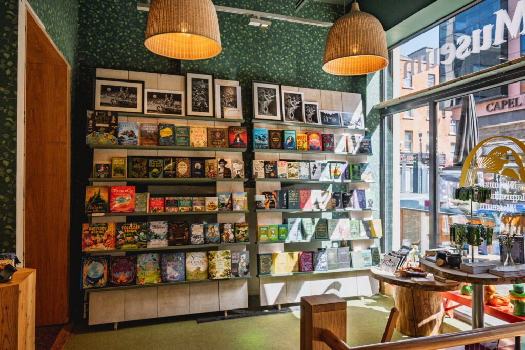 The museum gift shop, with sunlight shining through a large window. A bookshelf displaying books on irish folklore and mythology can be seen, along with tables displaying souvenirs and prints.