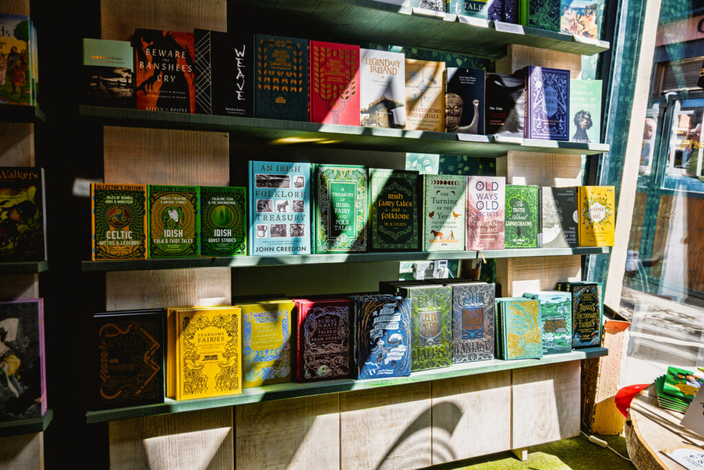 A close up of a gift shop bookshelf displaying books on Irish folklore and mythology. Sunlight illuminates some of the titles.