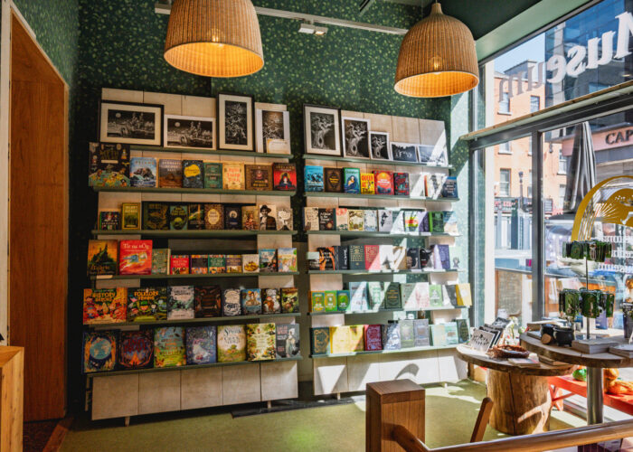 The museum gift shop, with sunlight shining through a large window. A bookshelf displaying books on irish folklore and mythology can be seen, along with tables displaying souvenirs and prints.